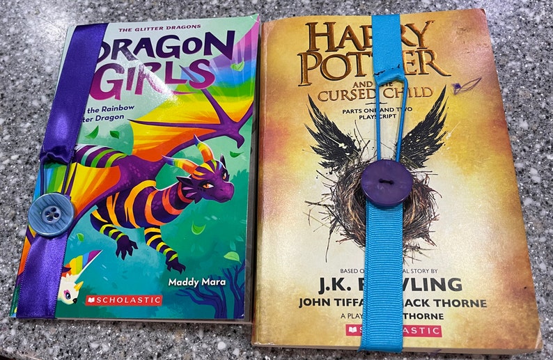 Peut inclure: Deux livres broch&eacute;s avec des couvertures color&eacute;es et des signets en ruban bleu. Le livre de gauche est intitul&eacute; "Dragon Girls" avec une illustration de dragon arc-en-ciel. Le livre de droite est intitul&eacute; "Harry Potter et l'Enfant Maudit" avec une illustration sombre d'un nid d'oiseau.