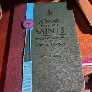 Peut inclure: Un livre reli&eacute; en cuir vert intitul&eacute; "A Year With The Saints: Daily Meditations With The Holy Ones Of God" par Paul Thigpen. Un marque-page en ruban bleu est attach&eacute; au livre avec un bouton violet.