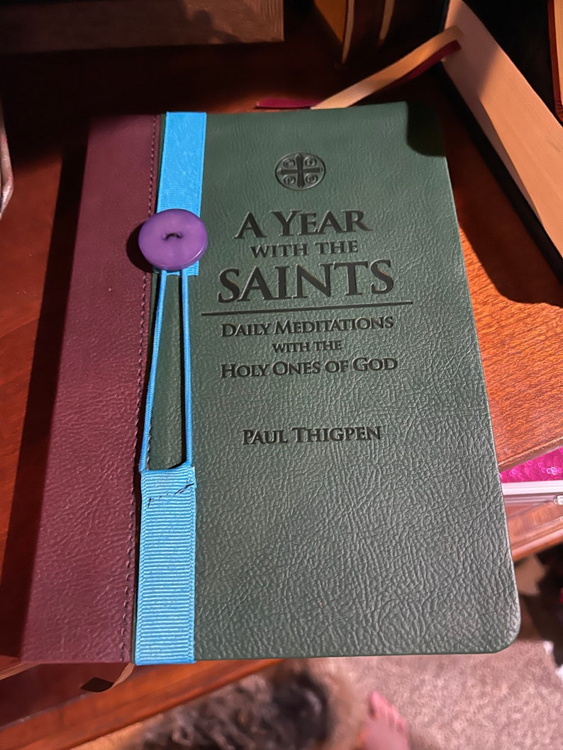 Peut inclure: Un livre reli&eacute; en cuir vert intitul&eacute; "A Year With The Saints: Daily Meditations With The Holy Ones Of God" par Paul Thigpen. Un marque-page en ruban bleu est attach&eacute; au livre avec un bouton violet.