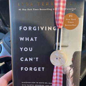 Peut inclure: Un livre reli&eacute; noir avec le titre "Forgiving What You Can't Forget" de Lysa TerKeurst. Le livre est un best-seller du New York Times et est marqu&eacute; "Uninvited". Un marque-page en ruban vichy rouge et blanc est attach&eacute; au livre.