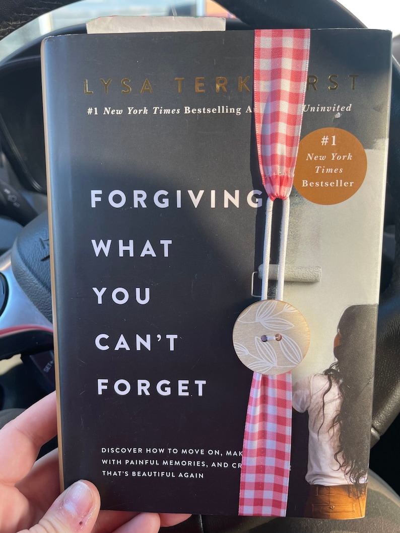 Peut inclure: Un livre reli&eacute; noir avec le titre "Forgiving What You Can't Forget" de Lysa TerKeurst. Le livre est un best-seller du New York Times et est marqu&eacute; "Uninvited". Un marque-page en ruban vichy rouge et blanc est attach&eacute; au livre.