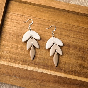 May include: A pair of dangle earrings with three tiers of beige and white ceramic leaves. The earrings are on a wooden surface.