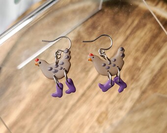 May include: A pair of whimsical earrings featuring a chicken design. The chickens are light brown with dark spots, red combs, and are wearing purple boots. The earrings have silver-colored hooks.