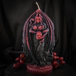 Pode incluir: Uma vela esculpida que retrata Lilith, uma figura da mitologia. A vela é principalmente preta e vermelha, apresentando uma mulher segurando uma caveira, com cobras e asas. A base da vela diz "LILITH".