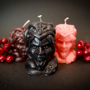 May include: Three sculpted candles in the shape of a demonic face, in black, brown, and peach colors. Red berries are placed on either side of the candles. The black candle is in the foreground, with a snake wrapped around the base.