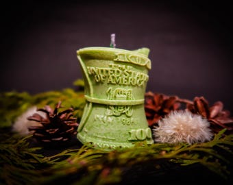 May include: A green candle shaped like a crumpled $100 bill. The candle has the words "THE UNITED STATES OF AMERICA" and "100" printed on it. The candle is sitting on a bed of green pine needles.