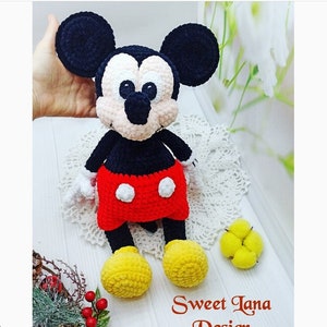 Puede incluir: Juguete de peluche Mickey Mouse de crochet con orejas negras, cuerpo negro, pantalones cortos rojos con lunares blancos y zapatos amarillos. El juguete está sentado en un encaje de crochet blanco.