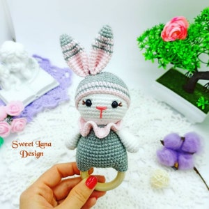 May include: A crocheted gray and pink bunny rattle with a wooden ring. The bunny has a pink nose and black eyes. The text "Sweet Lana Design" is visible in the image.