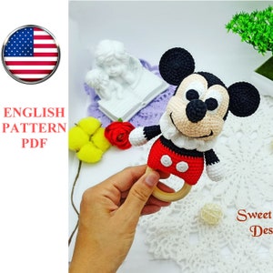 Op de afbeelding: Gehaakt Mickey Mouse rammelaar speelgoed met een houten ring. Het speelgoed is rood, zwart en wit met een glimlachend gezicht.