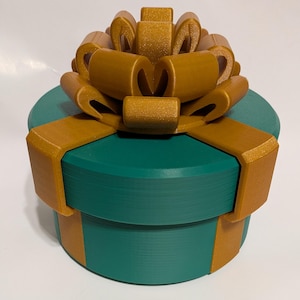 Puede incluir: Una caja de regalo redonda, de color verde azulado, con un lazo y una cinta dorados. El lazo es grande y adornado, con múltiples bucles. La cinta envuelve la caja. La caja es de color sólido, y el lazo y la cinta tienen un brillo metálico.