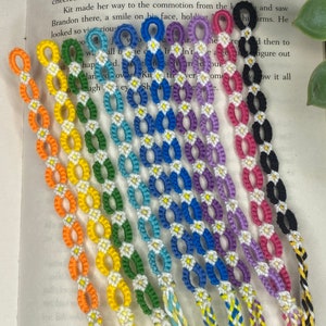 Könnte beinhalten: Ein Set mit 10 bunten Freundschaftsarmbändern mit weißen Gänseblümchen-Akzenten. Die Armbänder sind aus einem gewebten Schnurmuster gefertigt und in einem Regenbogen von Farben erhältlich, darunter Orange, Gelb, Grün, Blau, Lila, Pink und Schwarz.