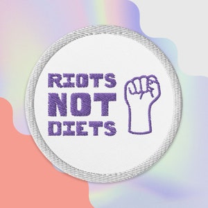 May include: A white round patch with a purple embroidered fist and the text "RIOTS NOT DIETS".