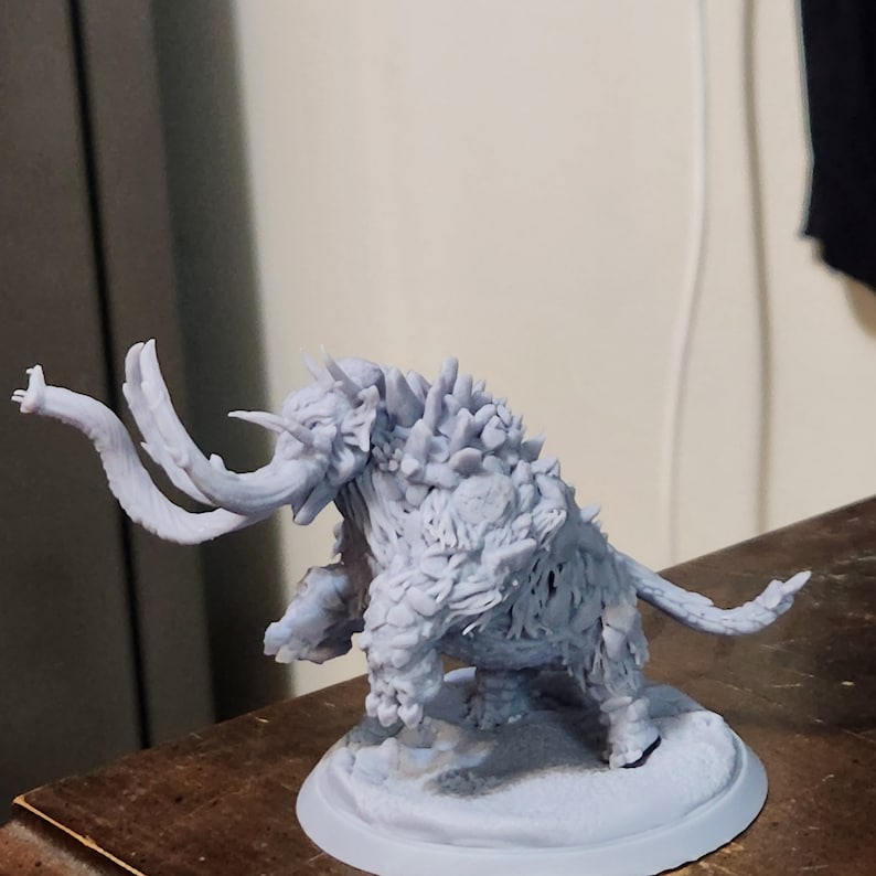 May include: A gray 3D printed miniature of a monstrous creature resembling a mammoth with a spiky back and tusks. The creature is standing on a round base.