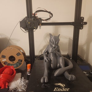 May include: A gray 3D printed figurine of a Pokemon character, Mewtwo, sitting with its arms crossed. The figurine is on a black 3D printer bed with the word "Ender" printed on it.