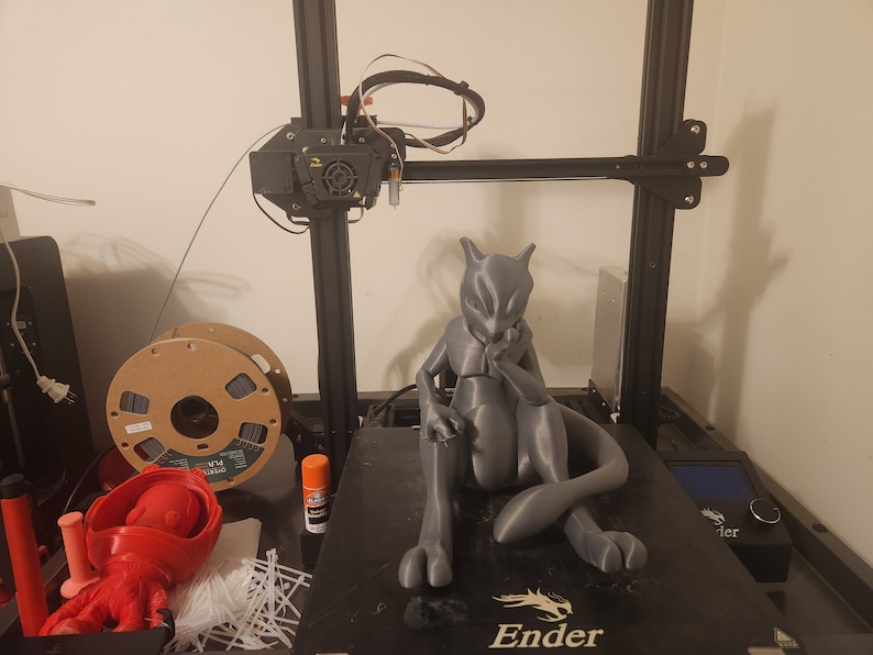May include: A gray 3D printed figurine of a Pokemon character, Mewtwo, sitting with its arms crossed. The figurine is on a black 3D printer bed with the word "Ender" printed on it.