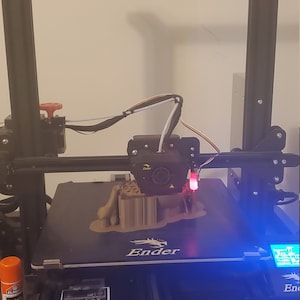 May include: A 3D printer with a brown plastic object being printed on the bed. The printer has a black frame and a blue light on the control panel. The text "Ender" is visible on the printer bed.