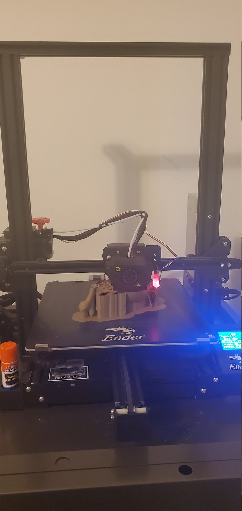 May include: A 3D printer with a brown plastic object being printed on the bed. The printer has a black frame and a blue light on the control panel. The text "Ender" is visible on the printer bed.