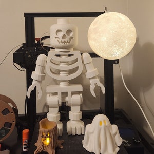 May include: A white 3D printed skeleton figure with a skull head, standing next to a white ghost and a small tree stump with a light inside. A white moon-shaped lamp hangs above the skeleton.