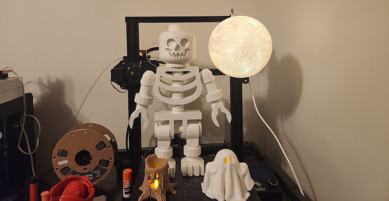 May include: A white 3D printed skeleton figure with a skull head, standing next to a white ghost and a small tree stump with a light inside. A white moon-shaped lamp hangs above the skeleton.