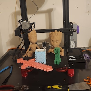 May include: A 3D printed model of a green Minecraft character, a pink Minecraft pickaxe, and two brown Groot figures from Guardians of the Galaxy. The figures are sitting on a black 3D printer bed with the word "Ender" printed on it.