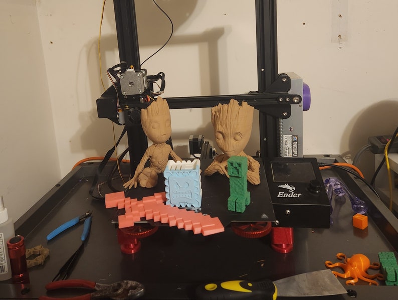 May include: A 3D printed model of a green Minecraft character, a pink Minecraft pickaxe, and two brown Groot figures from Guardians of the Galaxy. The figures are sitting on a black 3D printer bed with the word "Ender" printed on it.