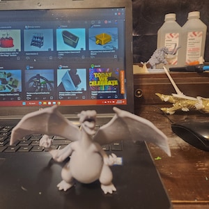 May include: A gray 3D printed dragon figurine with wings spread wide. The dragon is sitting on a black laptop keyboard. The laptop screen shows a website with several 3D printed designs.