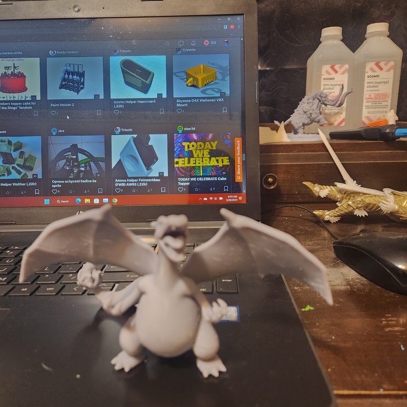 May include: A gray 3D printed dragon figurine with wings spread wide. The dragon is sitting on a black laptop keyboard. The laptop screen shows a website with several 3D printed designs.