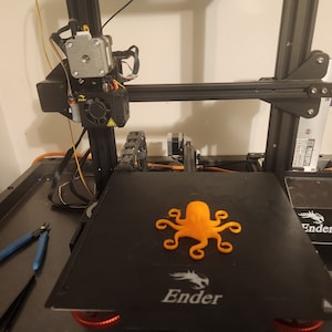 May include: An orange 3D printed octopus on a black 3D printer bed. The printer is a Creality Ender 3.