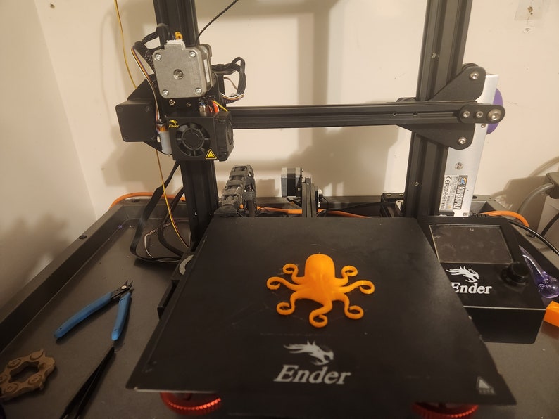 May include: An orange 3D printed octopus on a black 3D printer bed. The printer is a Creality Ender 3.
