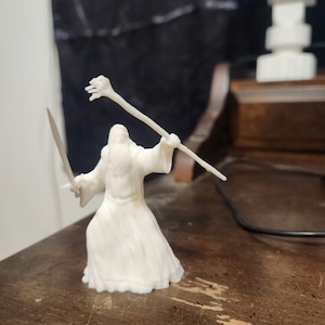 May include: White plastic figurine of a wizard holding a staff and a sword. The wizard is wearing a long robe and has a long beard.