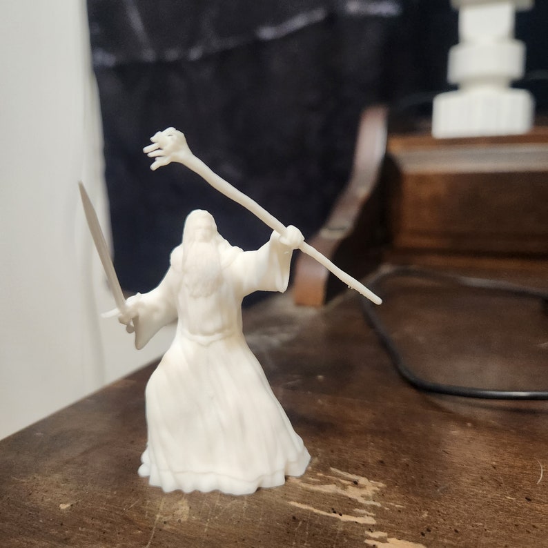 May include: White plastic figurine of a wizard holding a staff and a sword. The wizard is wearing a long robe and has a long beard.