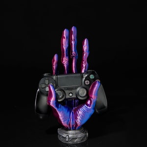May include: A black PlayStation controller held in a 3D printed hand with a purple and blue iridescent finish. The hand is on a black base.