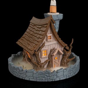 Könnte beinhalten: Ein beleuchtetes Miniatur-Spukhaus mit braunem Schindeldach, hellen Wänden und einem Steinkamin. Das Haus ist von einer Steinmauer und knorrigen Ästen umgeben. Die Fenster leuchten orange.