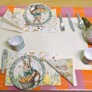 Puede incluir: Miniatura de juego de té con temática de Pascua con ilustraciones de conejos. Incluye platos, tazas, tetera, cubiertos y servilletas. Los platos presentan un conejo sosteniendo una cesta de zanahorias. El juego está dispuesto sobre una mesa.