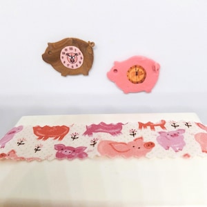 May include: Two pig-shaped clocks, one brown and one pink, sit above a white surface with a pig-themed pattern. The clocks have clock faces. The pattern on the surface features pink pigs, flowers, and other farm-related elements.