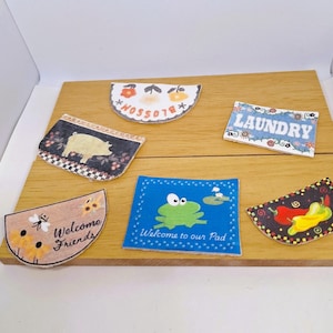 May include: A collection of miniature decorative mats with various designs. One mat has the word "Laundry", another says "Welcome Friends", and a third says "Welcome to our Pad". Other mats feature a pig, flowers, and a frog.