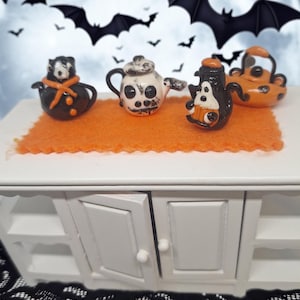 Pode incluir: Um conjunto de quatro bules em miniatura com tema de Halloween nas cores preto, branco e laranja. A coleção inclui um bule preto com um cachecol laranja, um bule branco em forma de caveira, um bule preto com um fantasma e um bule laranja com bolinhas pretas.