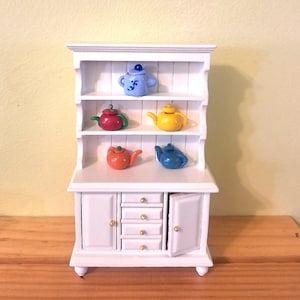 Op de afbeelding: Een witte miniatuurkast met planken met zes kleurrijke theepotten. De theepotten zijn in verschillende kleuren, waaronder blauw, rood, oranje en geel. De kast heeft een gesloten deur en lades met goudkleurige knoppen.