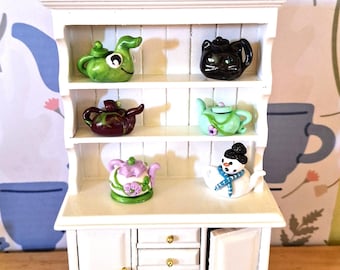 May include: A white miniature cabinet with multiple shelves displaying a collection of decorative teapots. The teapots are various colors and designs, including a green frog, a black cat, and a snowman. The cabinet has drawers and doors with gold knobs.