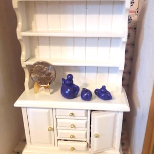 Pode incluir: Miniatura de aparador branco com prateleiras, gavetas e portas de armário. Inclui uma pequena chaleira de cerâmica azul, açucareiro e cremeira. Uma moeda é exibida em um suporte dourado. O aparador fica em uma superfície estampada.