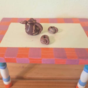 Pode incluir: Uma mesa em miniatura pintada à mão com um padrão xadrez rosa e laranja. Apresenta uma pequena chaleira marrom e dois pequenos objetos redondos. As pernas da mesa são pintadas de branco, azul e laranja.