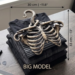 Op de afbeelding: Een stapel opgevouwen zwarte shirts met een decoratieve hanger met skeletthema. De hanger is ongeveer 30 cm breed en 33 cm hoog. De afbeelding bevat de tekst "DIMENSIONS GUIDE" en "BIG MODEL".