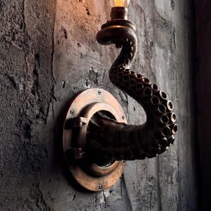 Op de afbeelding: Bronzen wandlamp met een octopus tentakel ontwerp. De tentakel buigt omhoog en houdt een vintage stijl gloeilamp vast. De armatuur is gemonteerd op een getextureerde, donkergrijze muur.