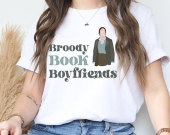 May include: White t-shirt with a graphic of a man in a blue shirt and brown jacket. The text "Broody Book Boyfriends" is printed in black and gray letters.