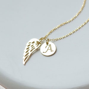 May include: A gold necklace with a delicate chain and a pendant featuring a small angel wing and a round charm with the letter 'A' engraved on it.