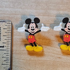 May include: Two plastic Mickey Mouse figures with outstretched arms and yellow shoes. The figures are approximately 1 inch tall.
