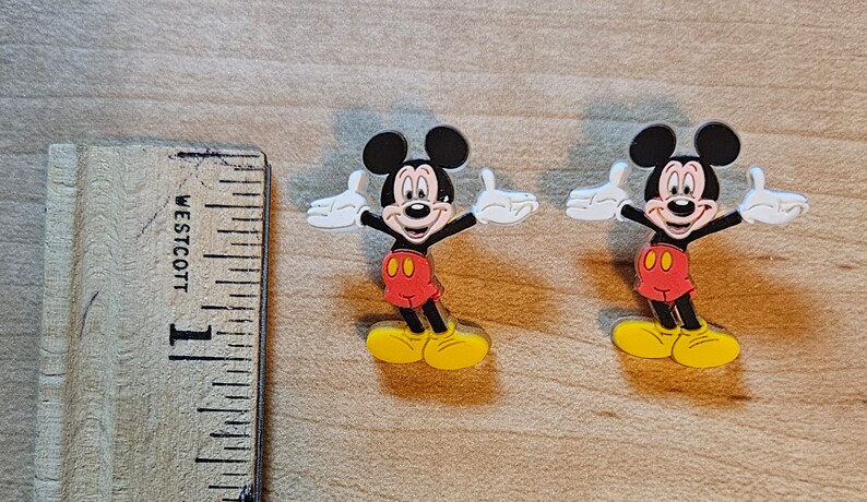 May include: Two plastic Mickey Mouse figures with outstretched arms and yellow shoes. The figures are approximately 1 inch tall.