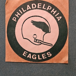 May include: A brown patch with a black circle containing the words "Philadelphia Eagles" in white lettering. Inside the circle is a pink outline of a football helmet with a wing on top.