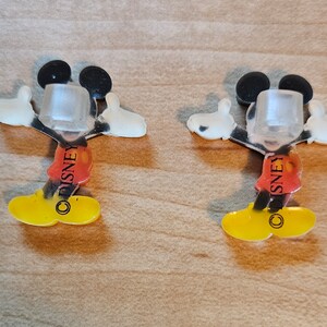May include: Two clear plastic Mickey Mouse figures with red shorts and yellow shoes. The figures have the word "DISNEY" printed on their shorts.