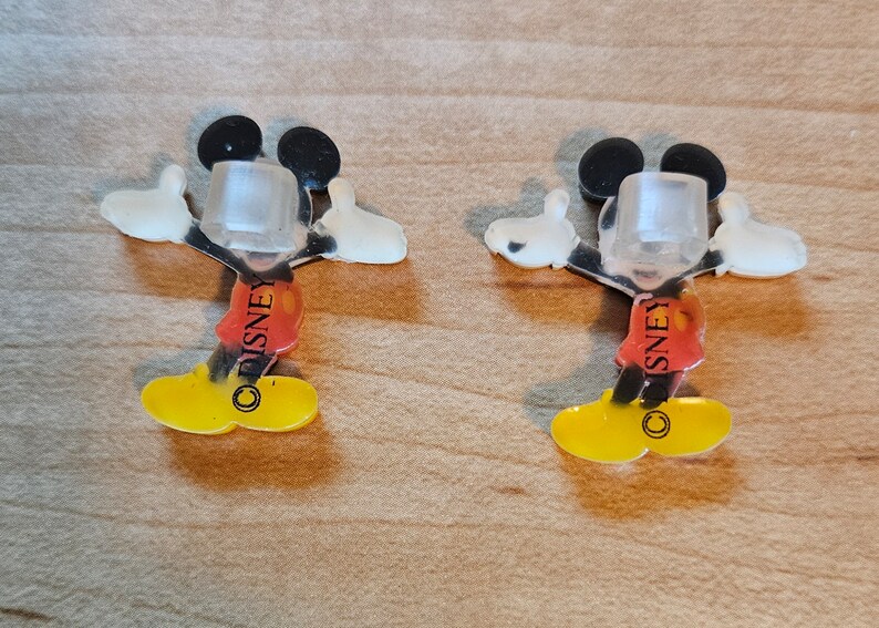 May include: Two clear plastic Mickey Mouse figures with red shorts and yellow shoes. The figures have the word "DISNEY" printed on their shorts.